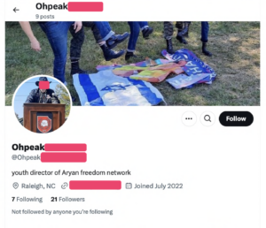 X (formerly Twitter) profile for Ohpeak. full userid, website, and person's face is redacted. profile pic shows ohpeak in black hat and black jacket standing outside behind a podium with the aryan freedom network logo on the front. banner image shows skinhead boots as if the are going to stomp on three flags celebrating pride, the state of israel, and harris for president. his bio reads "youth director of Aryan freedom network", location is Raleigh, NC, Joined date is July 2022. He has 21 followers and is following 7.