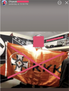 ohpeak in what appears to be his bedroom, posing while holding a ku klux klan flag. behind him is a collection of various white power and nazi flags and memorabilia.